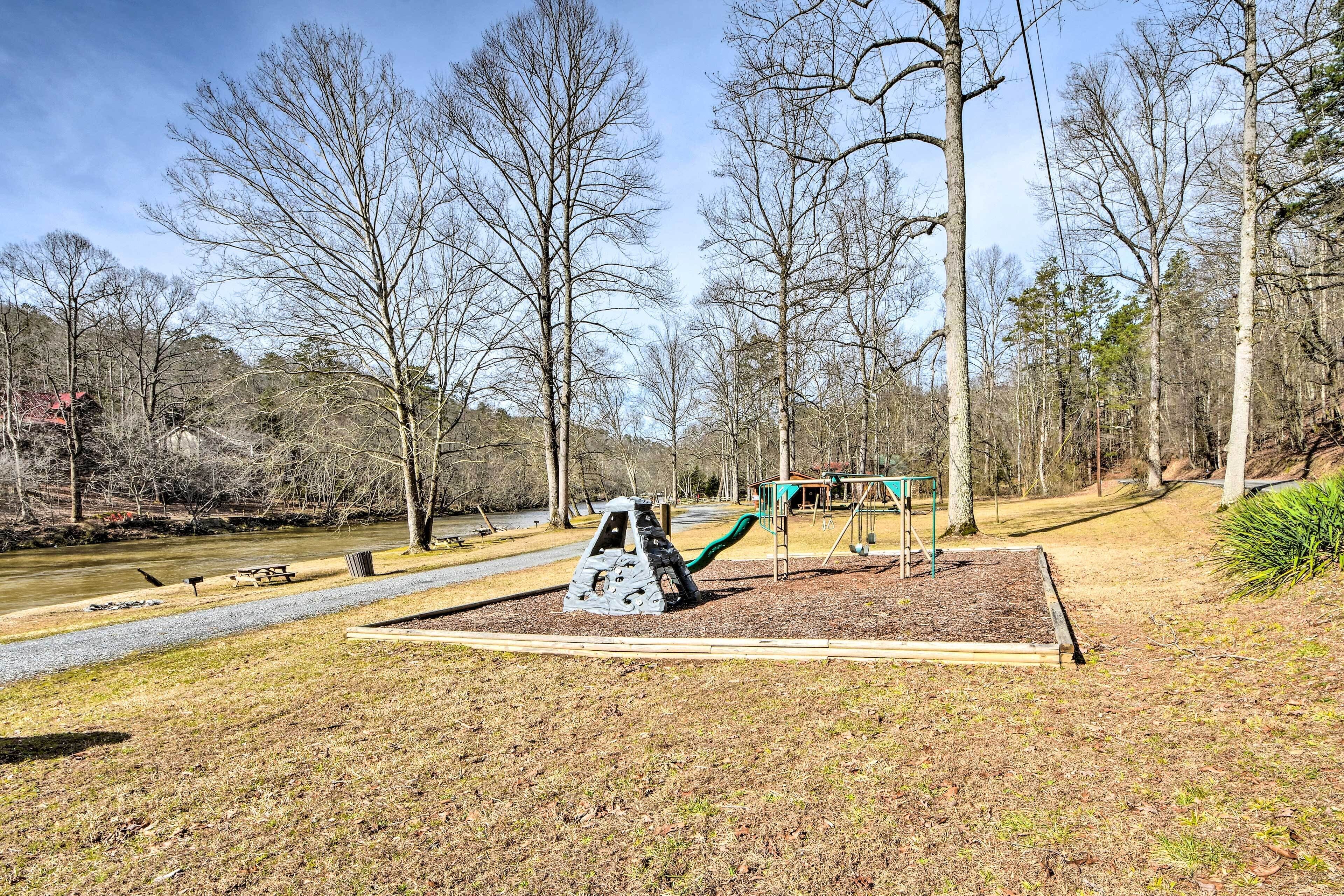 A playground with a swing set and a climbing structure, surrounded by trees and a path.
