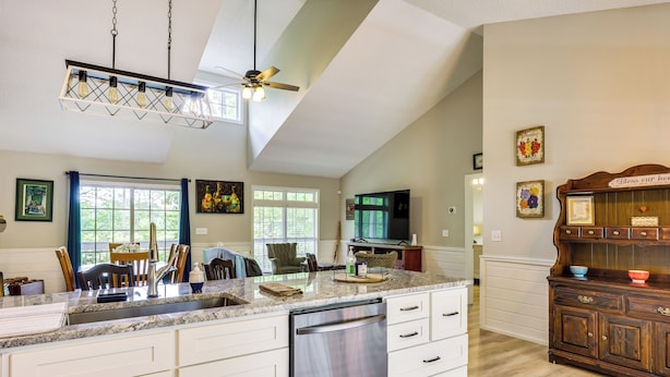 A kitchen with a dishwasher, a large island with a sink, and a ceiling fan. There are framed pictures on the wall and a wooden cabinet with a sign above it. - Ellijay Escape: Hike, Shop, Explore & More! (Ellijay)