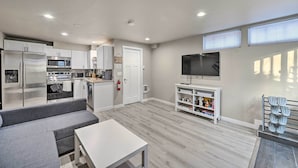 A modern kitchen with stainless steel appliances, a grey sofa, and a flat-screen TV on a white stand.