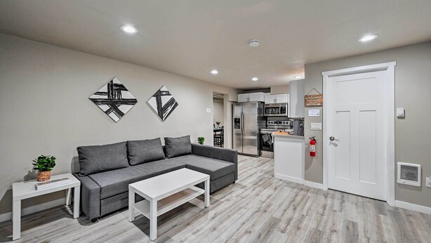 A modern living room with a grey sectional sofa, a white coffee table, and a small side table with a plant. There is a kitchen area in the background with stainless steel appliances.