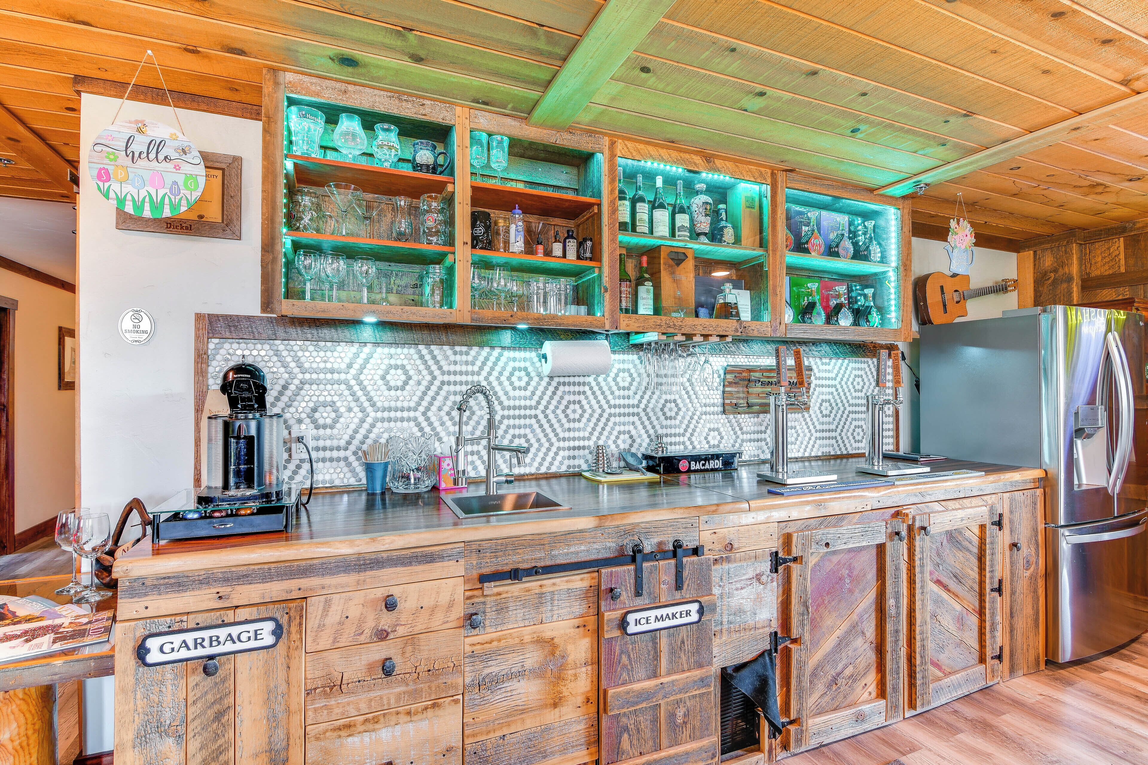 A rustic kitchen with wooden cabinets, a stainless steel refrigerator, and a bar area with shelves of bottles and glasses.