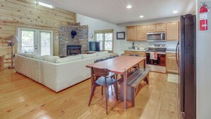 A spacious living room with a wooden floor, a sofa, a dining table with chairs, a stone fireplace, and a kitchen area with wooden cabinets and appliances. - Coosawattee River Resort Home Mins From Blue Ridge (Ellijay)
