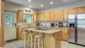 A kitchen with wooden cabinets, a stainless steel refrigerator, a granite countertop, and three bar stools. - Murrayville Lakefront Cabin w/ Boat Slip & Grill! (Murrayville)