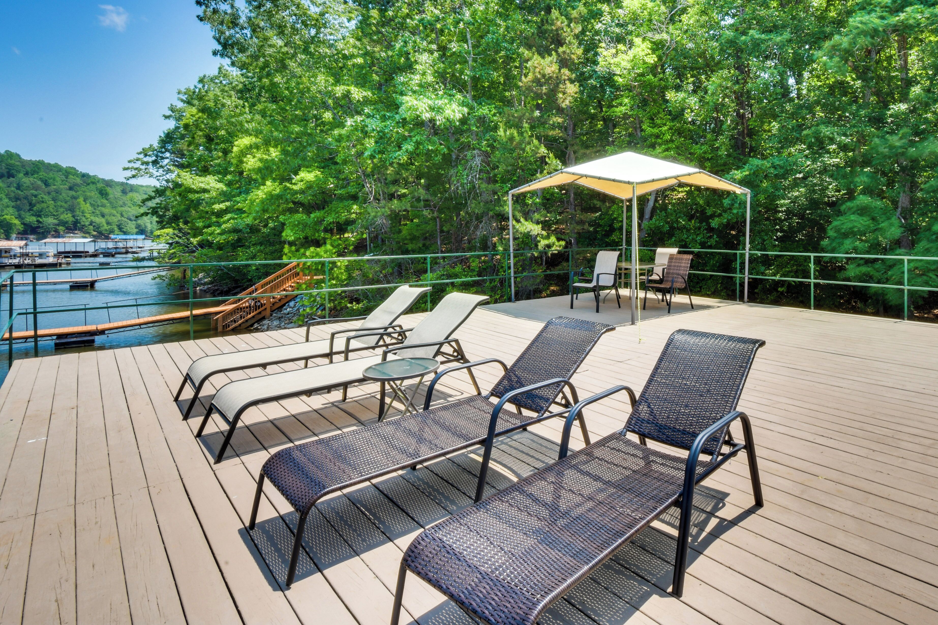 Une terrasse en bois avec des chaises en métal et un espace abrité, offrant une vue sur une étendue d’eau et entourée d’arbres.