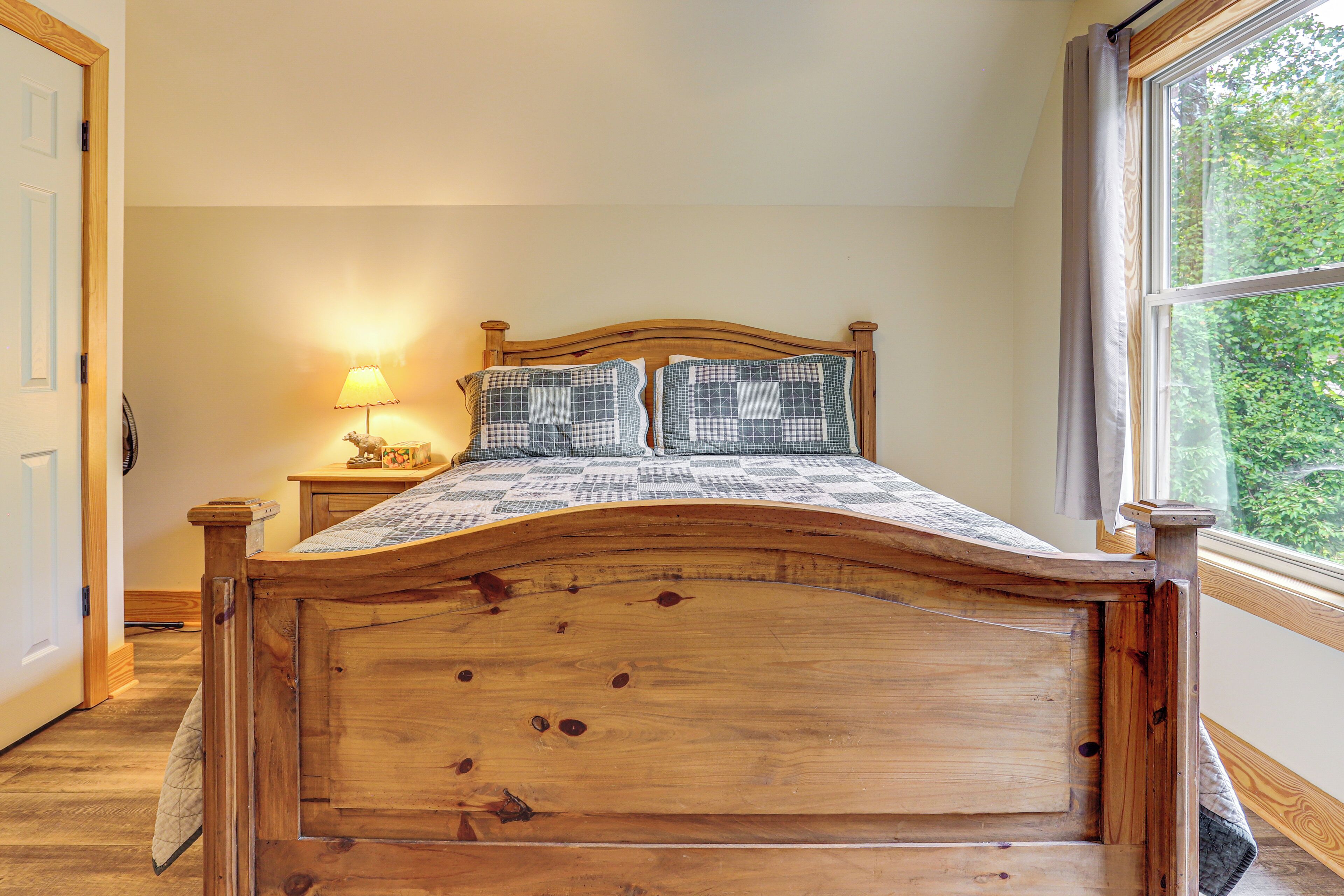 A wooden bed with a headboard, a bedside table with a lamp, and a window with a view of greenery.