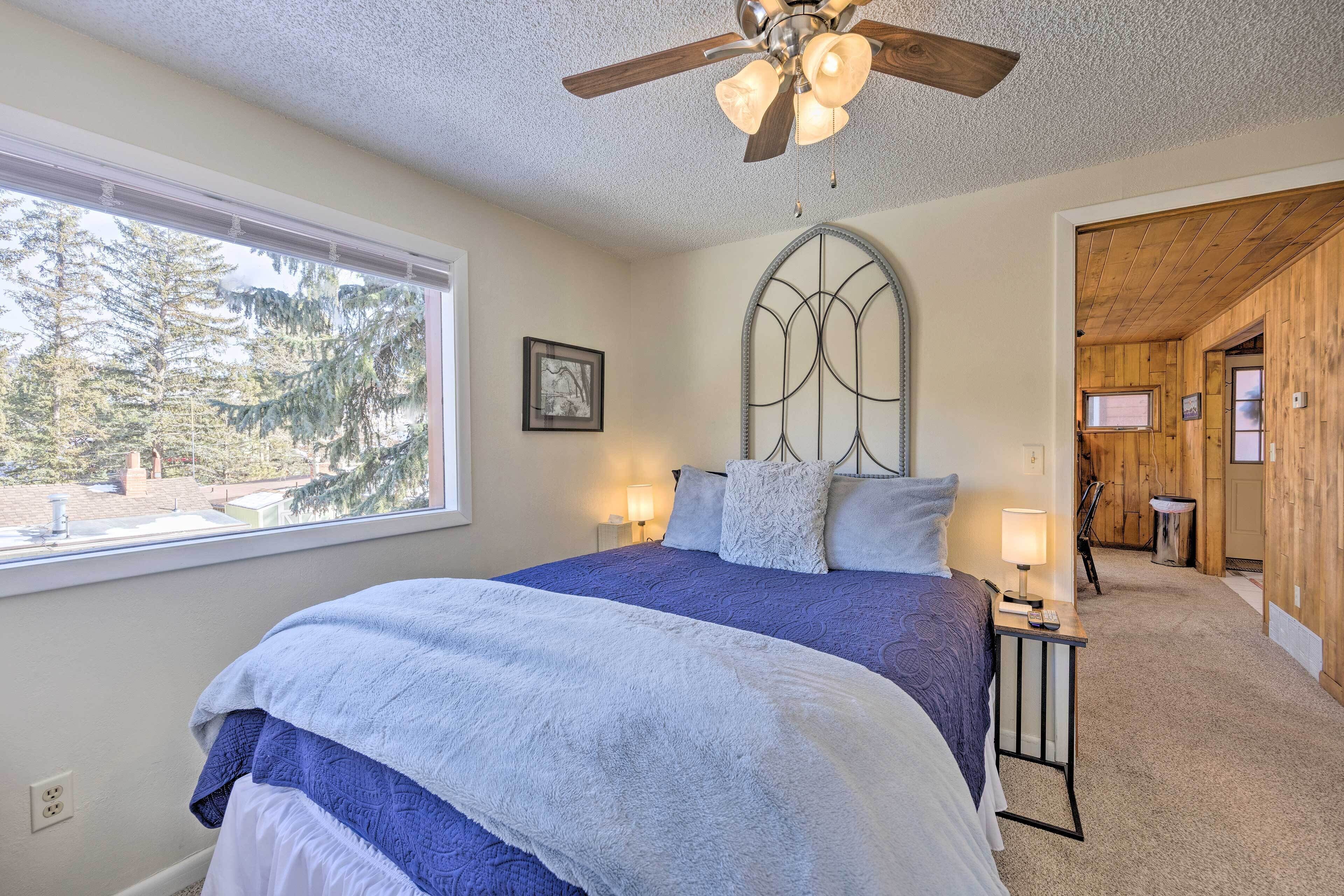 A bedroom with a large bed, a ceiling fan, a window with a view of trees, and a wooden wall.