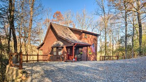 A rustic wooden cabin with a porch, surrounded by trees and gravel.
