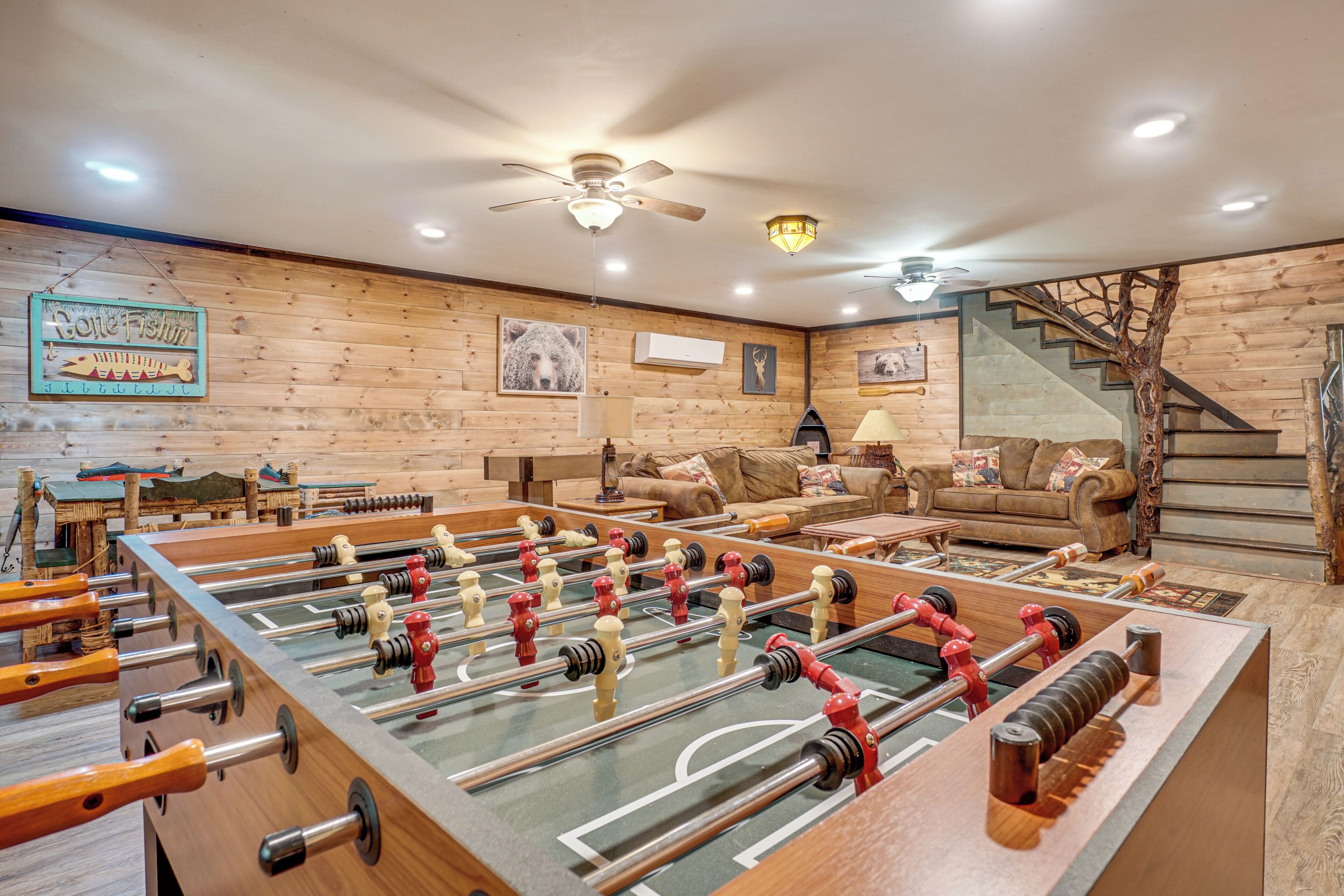 A room with a foosball table, wooden walls, a sofa, a staircase, and a ceiling fan.