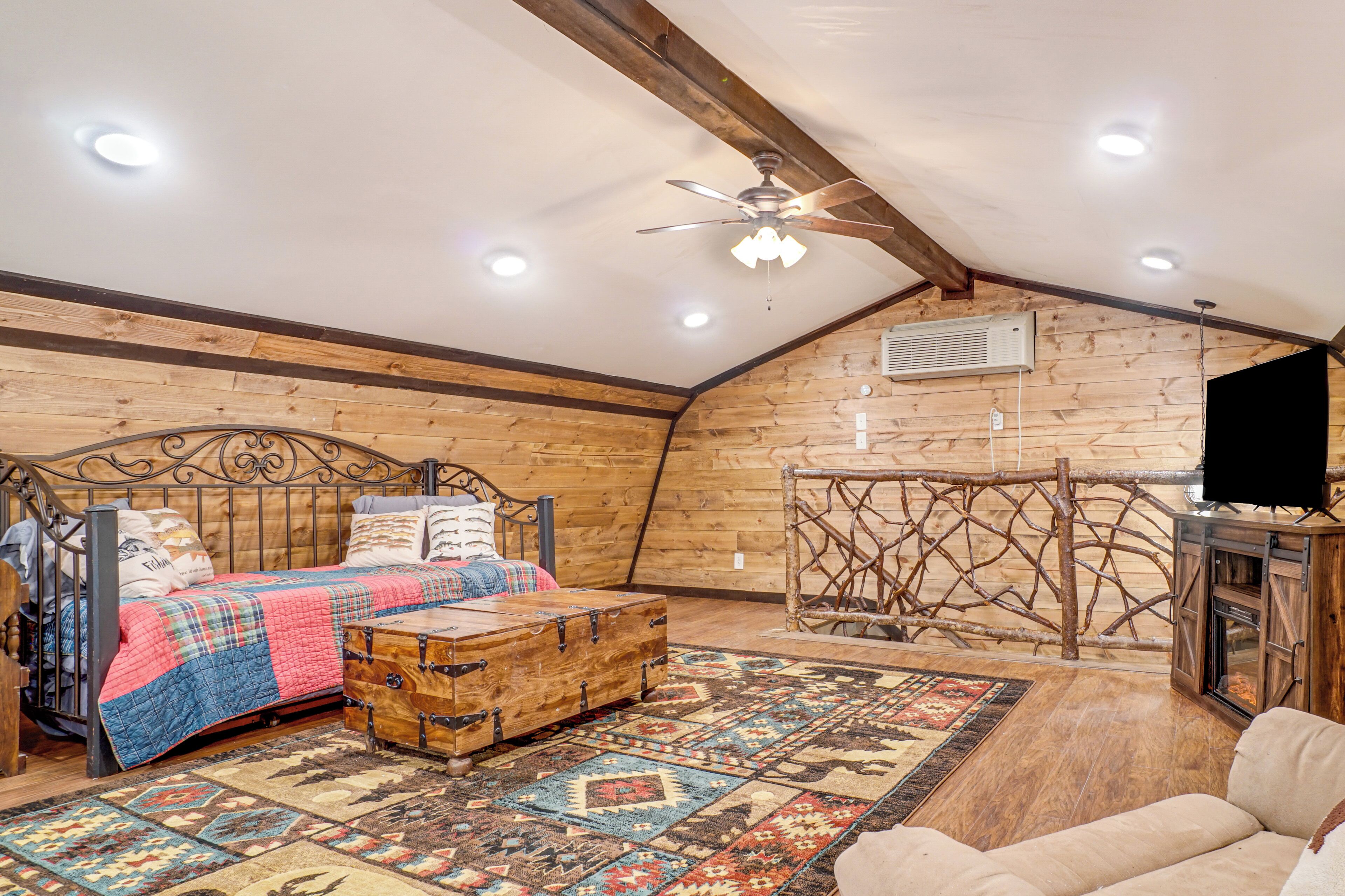 A cozy loft bedroom with a wooden ceiling, a bed with a plaid quilt, a wooden chest, and a rustic TV stand.