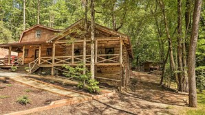 A rustic log cabin with a porch and a covered deck, surrounded by a forested area.