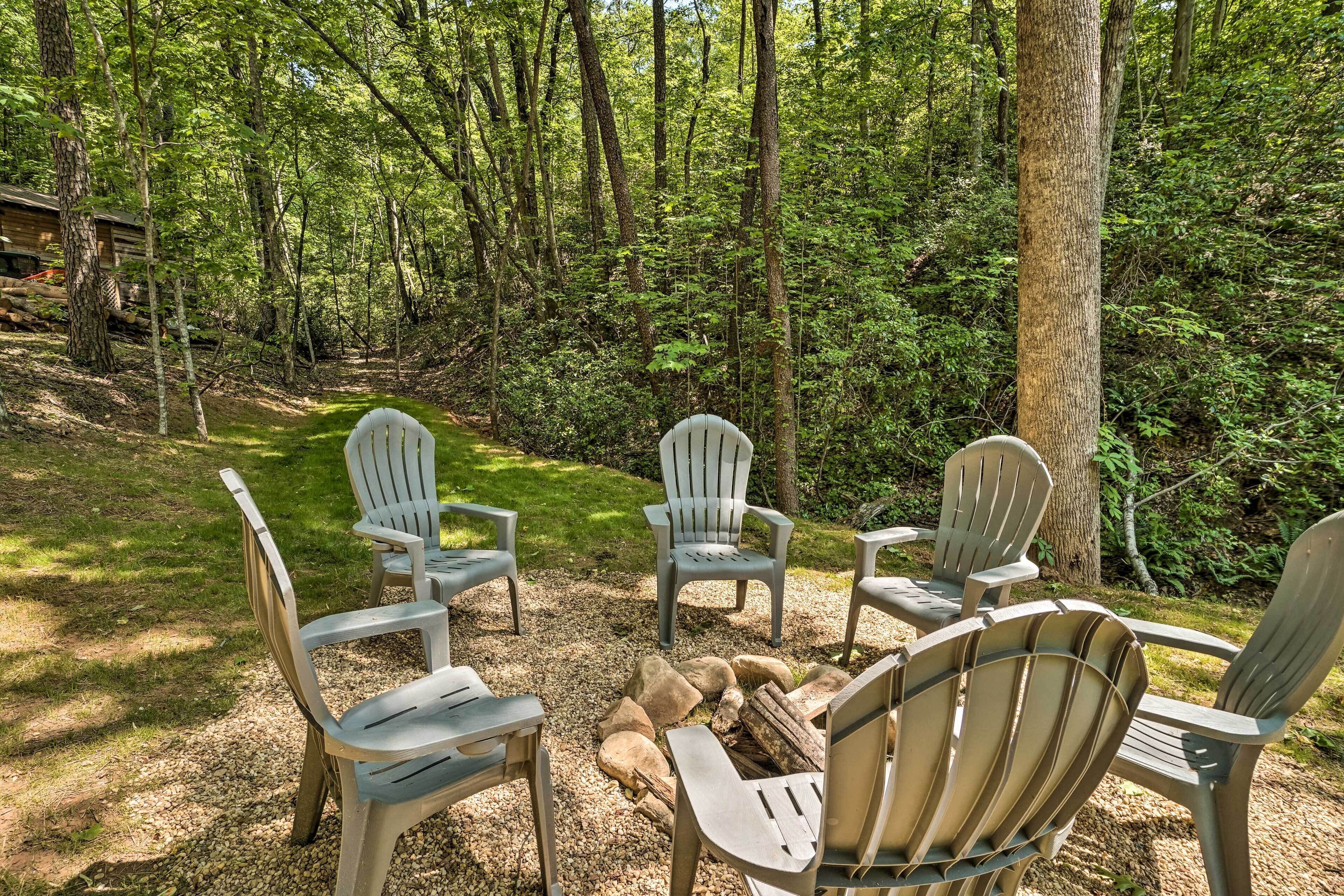 A forested area with Adirondack chairs arranged around a fire pit.