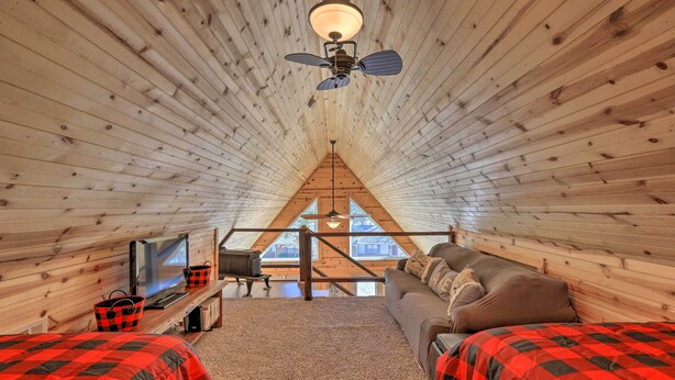 Un dormitorio acogedor en el ático con techo inclinado, ventilador de techo, televisor, sofá y vistas al exterior a través de una ventana.