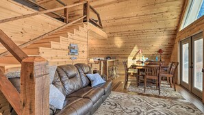Un acogedor interior de cabaña con sofá de cuero, escalera de madera, zona de comedor y ventanas amplias.