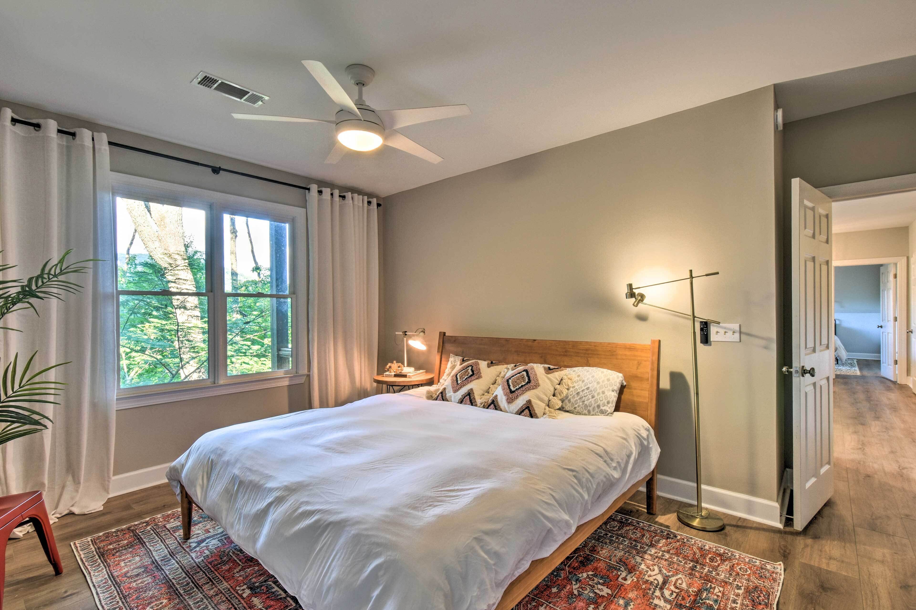 A bedroom with a bed, a ceiling fan, a nightstand with a lamp, a window with curtains, and a door leading to another room.