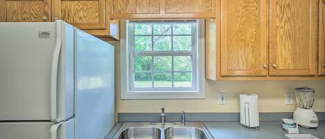 Una cocina con armarios de madera, un lavamanos de acero inoxidable, un refrigerador, una ventana y una licuadora.