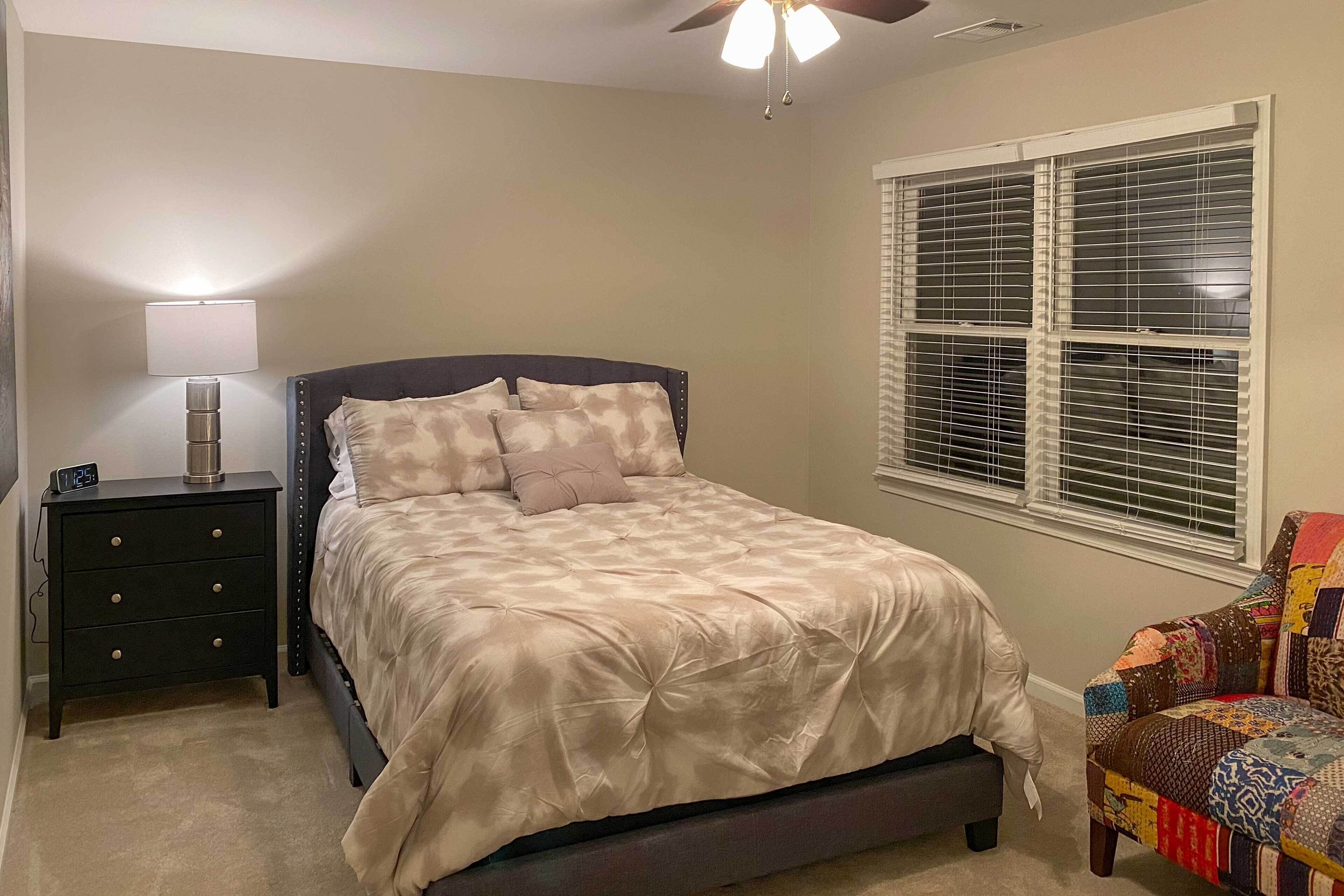 A bedroom with a bed, a nightstand, a lamp, a ceiling fan, a window with blinds, and a colorful chair.
