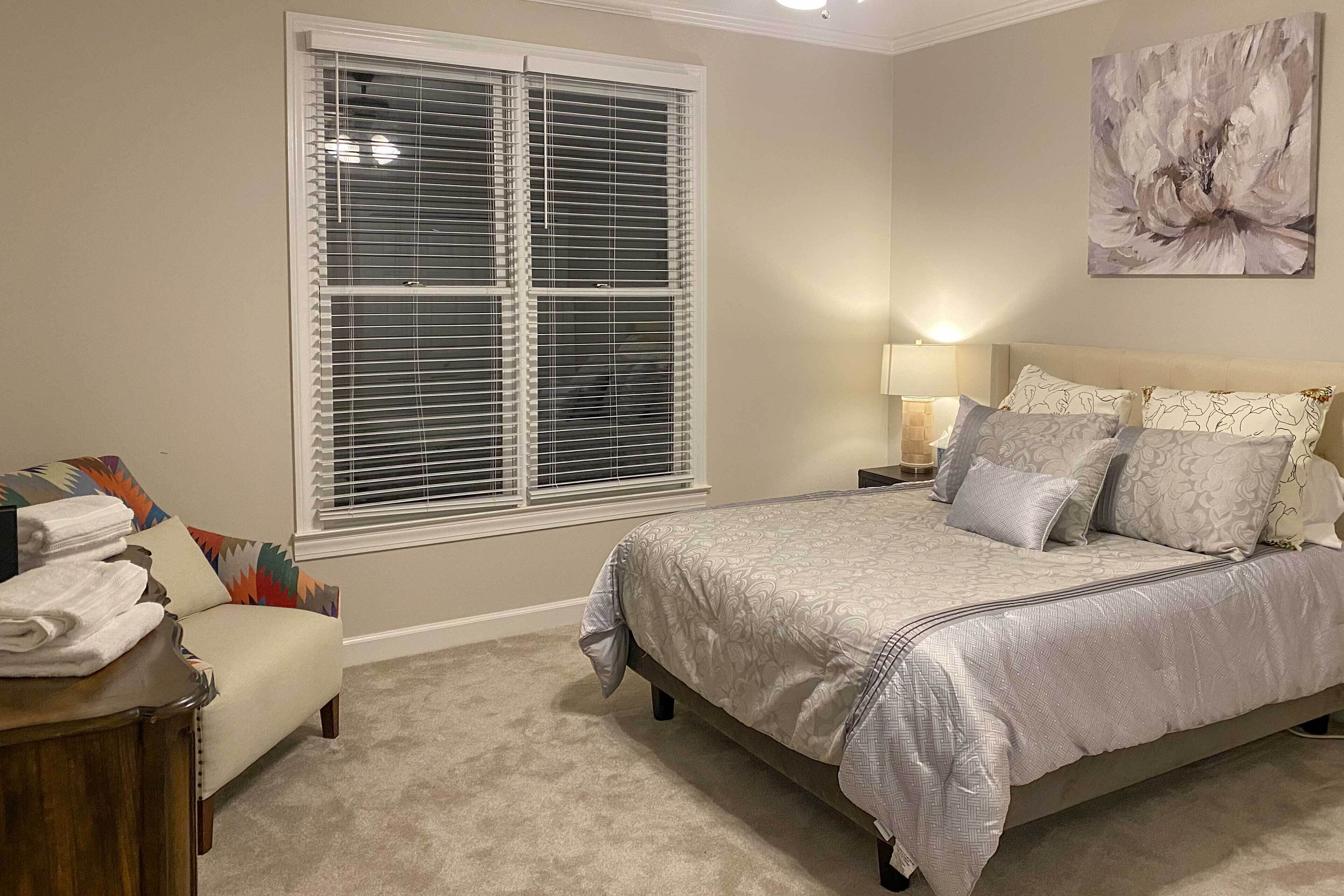 A bedroom with a large bed, a nightstand, a lamp, a chair, and a window with blinds.