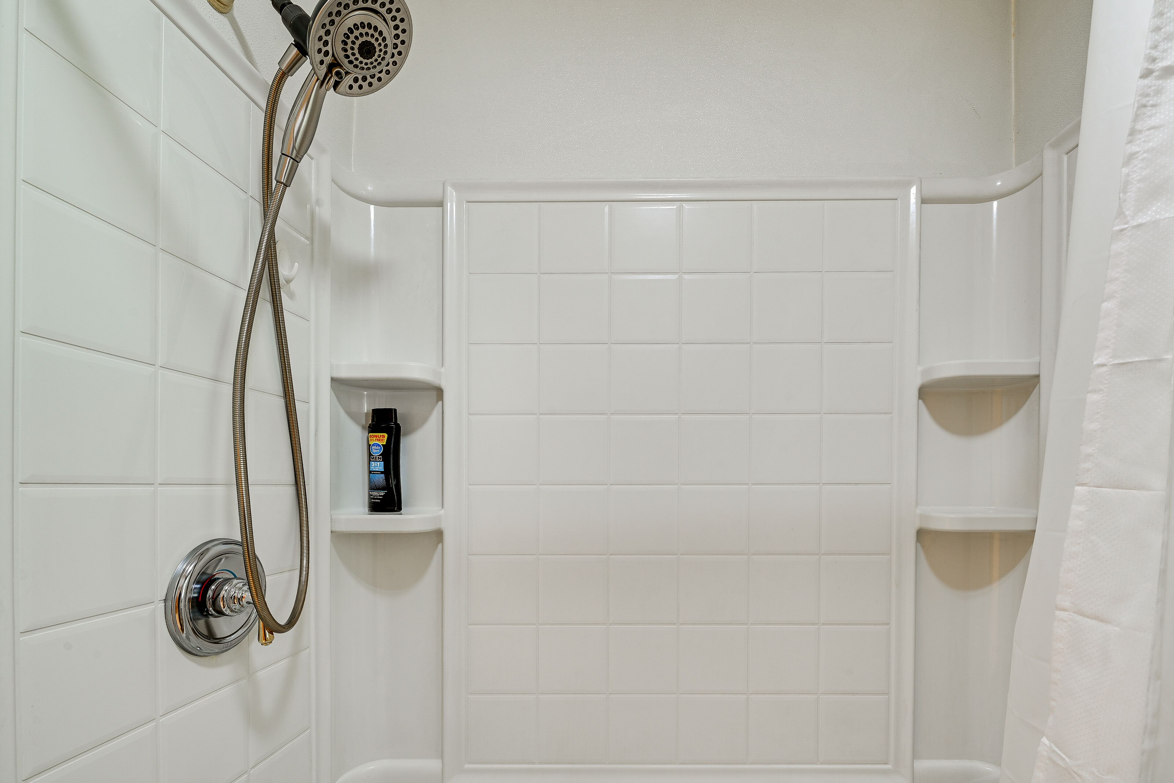A bathroom with white tiled walls, a shower with a handheld showerhead, and a shelf with toiletries.