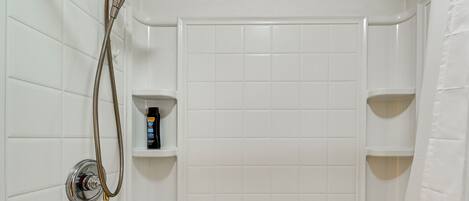 A bathroom with white tiled walls, a shower with a handheld showerhead, and a shelf with toiletries.