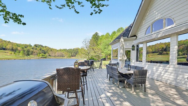 Une terrasse dotée de mobilier d’extérieur et d’un barbecue, offrant une vue sur un lac et entourée d’arbres.