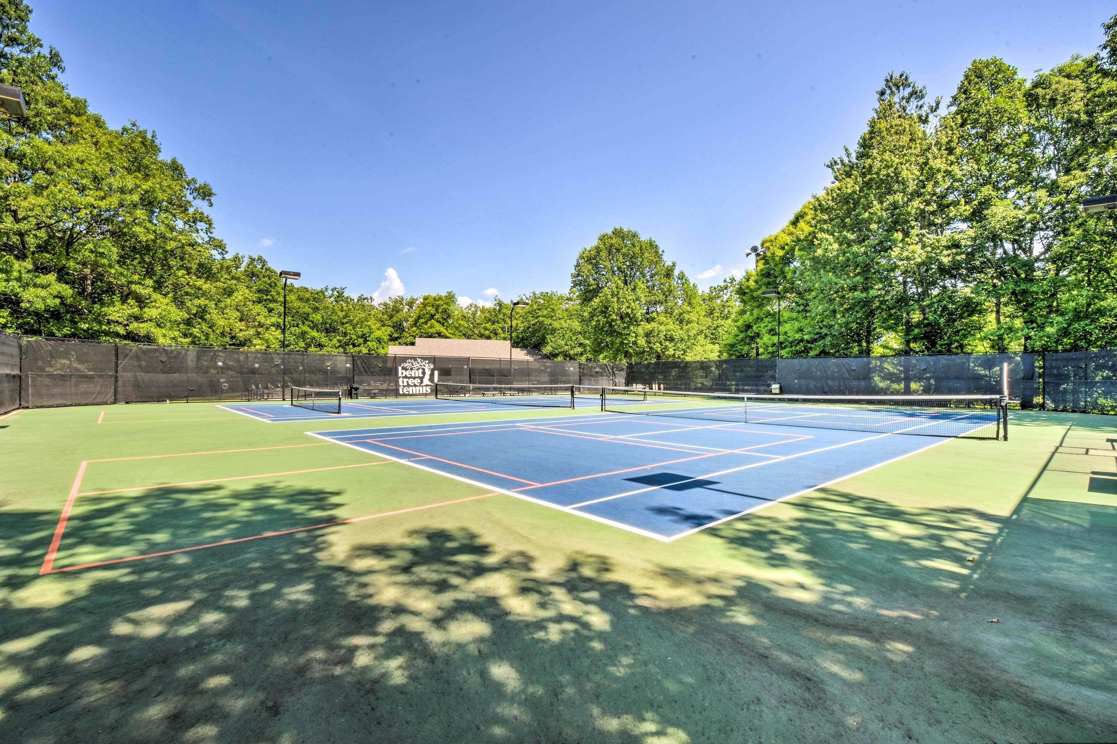 Un court de tennis en plein air, doté d’un filet, entouré d’arbres et délimité par une clôture.