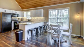 A kitchen with a wooden ceiling, white cabinets, a black refrigerator, a microwave, and a dining area with white chairs and stools. - Sparta Home w/ On-site Lake Access! (Milledgeville)