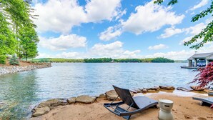 A lakeside view with a lounge chair, a small table, and a rustic cabin in the background. - Waterfront Escape on Blue Ridge Lake w/ Dock! (Morganton)