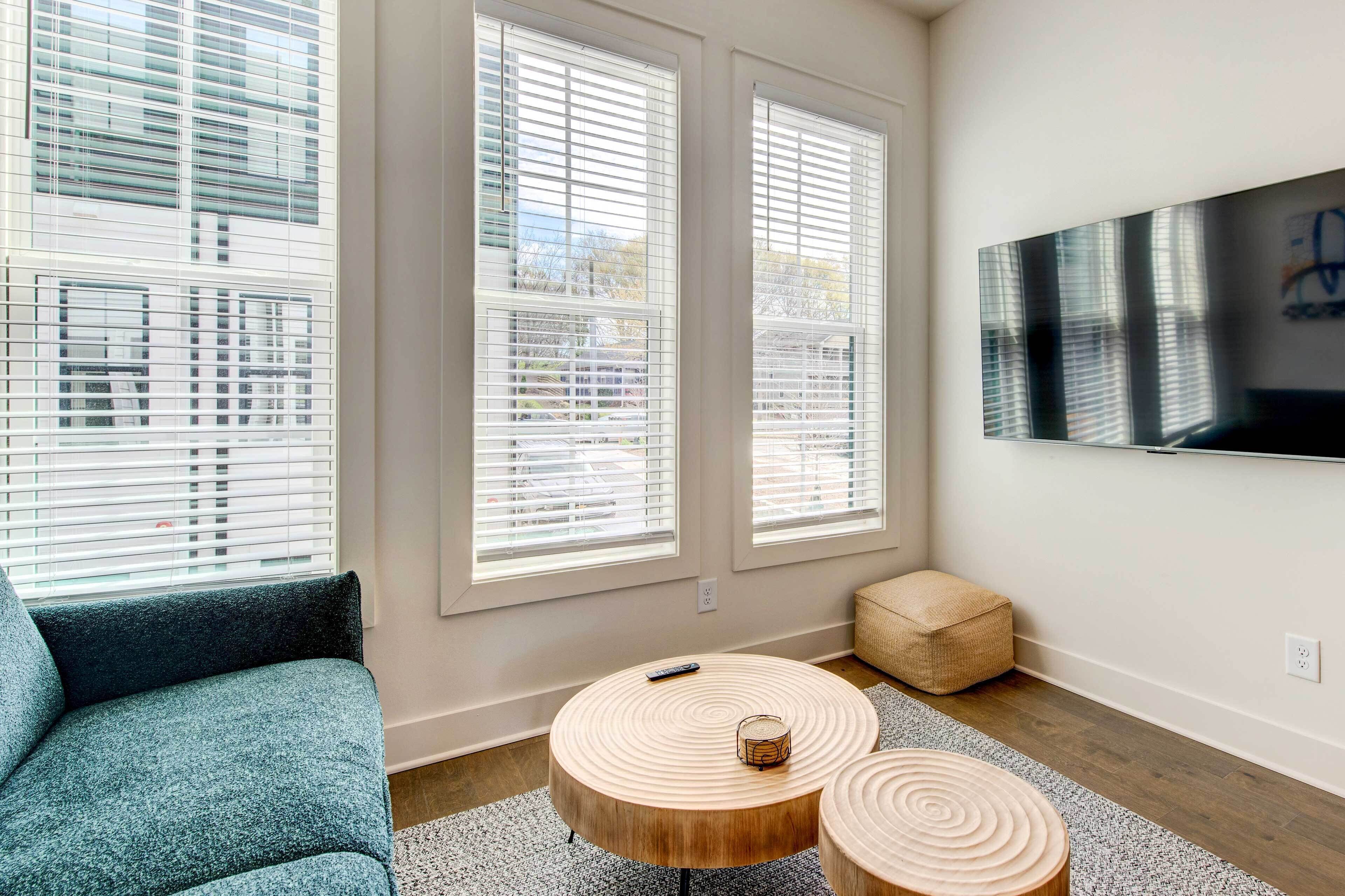 A modern living room with a sofa, two round wooden coffee tables, a TV mounted on the wall, and large windows with blinds.