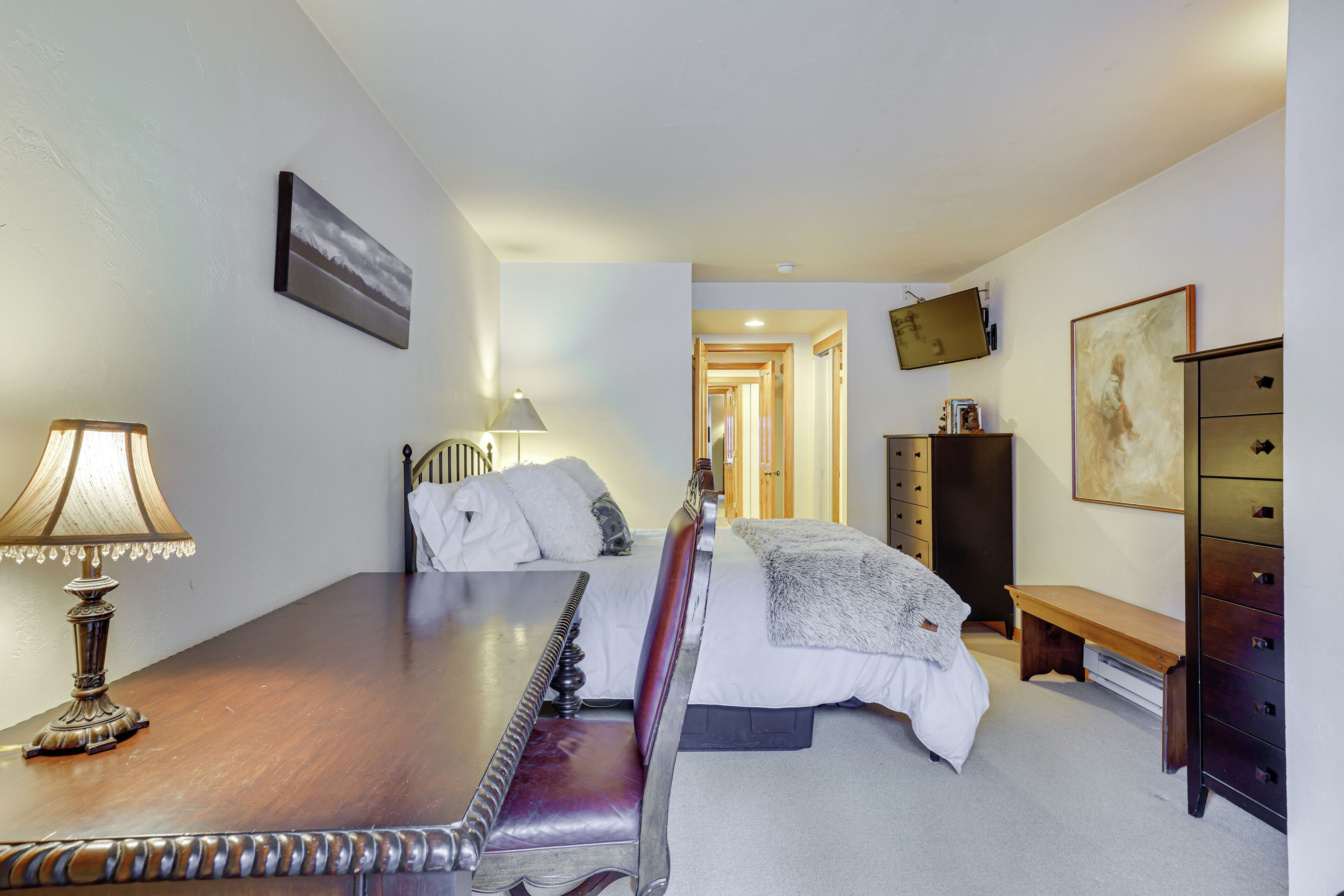 A hotel room with a wooden desk, a bed with white linens, a bench, a dresser, and a television on the wall.