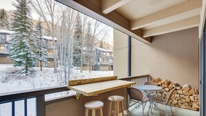 Un balcon avec une table en bois et deux tabourets, une pile de bois de chauffage, et une vue sur des arbres et des bâtiments recouverts de neige.