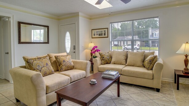 A living room with two beige sofas, a wooden coffee table, a large window with blinds, a ceiling fan, and a framed picture on the wall.