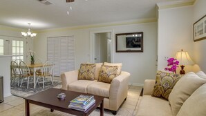 A living room with a beige sofa, a wooden coffee table with magazines, a dining area with a white table and chairs, and a kitchen visible in the background.