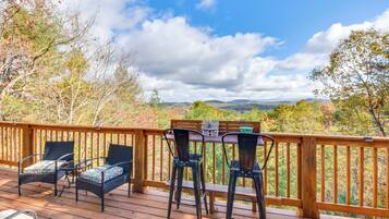 A wooden deck with wicker chairs and a table, overlooking a scenic landscape with trees and hills.