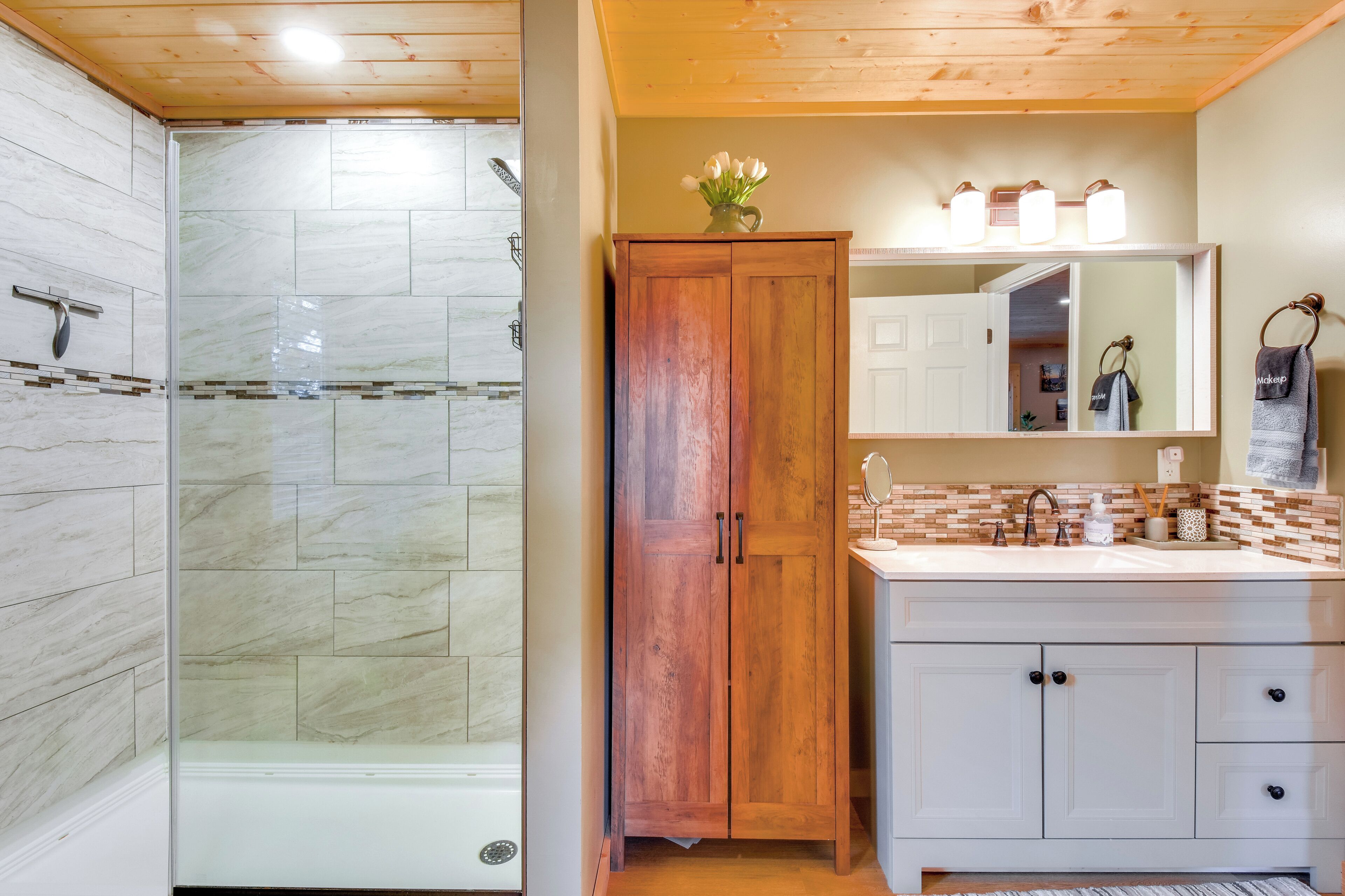 A bathroom with a glass-enclosed shower, wooden cabinets, a white vanity with a mirror, and a tiled backsplash.