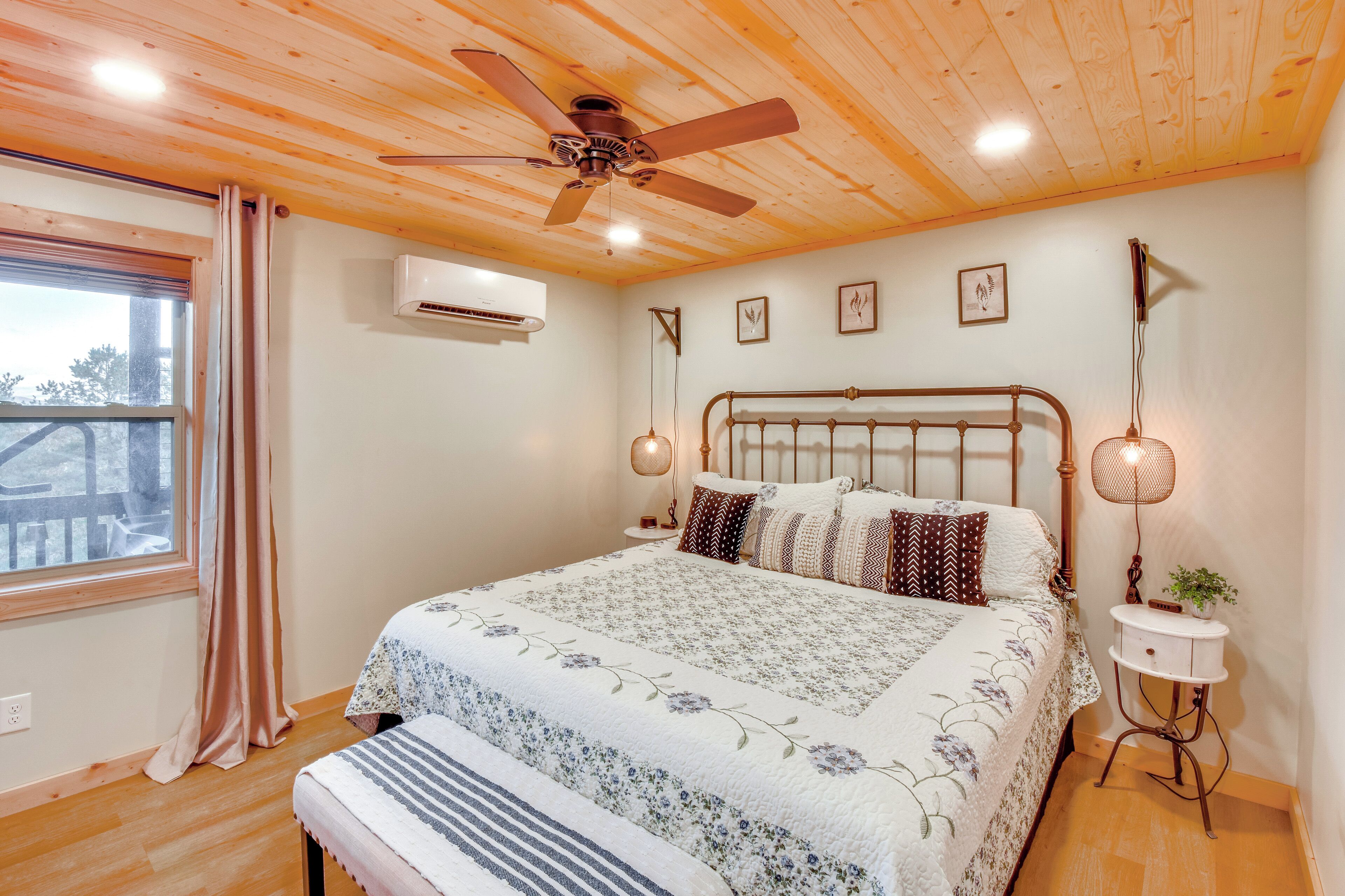 A bedroom with a wooden ceiling, a bed with floral bedding, a bedside table with a lamp, and a window with curtains.