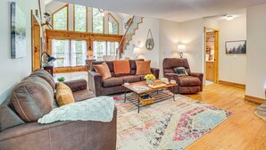 A spacious living room with a brown leather sofa, a brown armchair, and a wooden coffee table. There is a staircase leading to an upper level, and a large window with a view of trees. - North Georgia Cabin w/ Hot Tub & Community Perks! (Jasper)