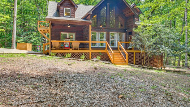 A log cabin with a wooden deck, a staircase, and a large window overlooking a forested area. - North Georgia Cabin w/ Hot Tub & Community Perks! (Jasper)