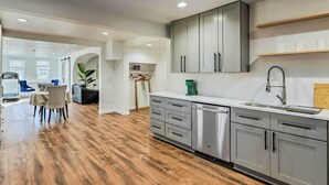 A modern kitchen with grey cabinets, a stainless steel dishwasher, and a wooden dining area with a table and chairs.