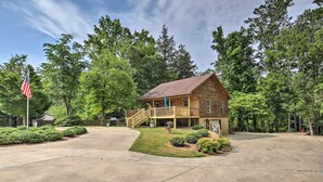 A two-story wooden cabin with a deck and a covered porch, surrounded by lush greenery and a clear sky.