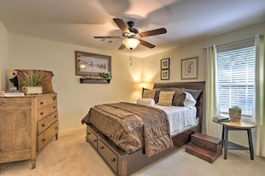 Une chambre à coucher comprenant un lit, une commode, un ventilateur de plafond, une petite table avec une plante et une fenêtre avec des stores.