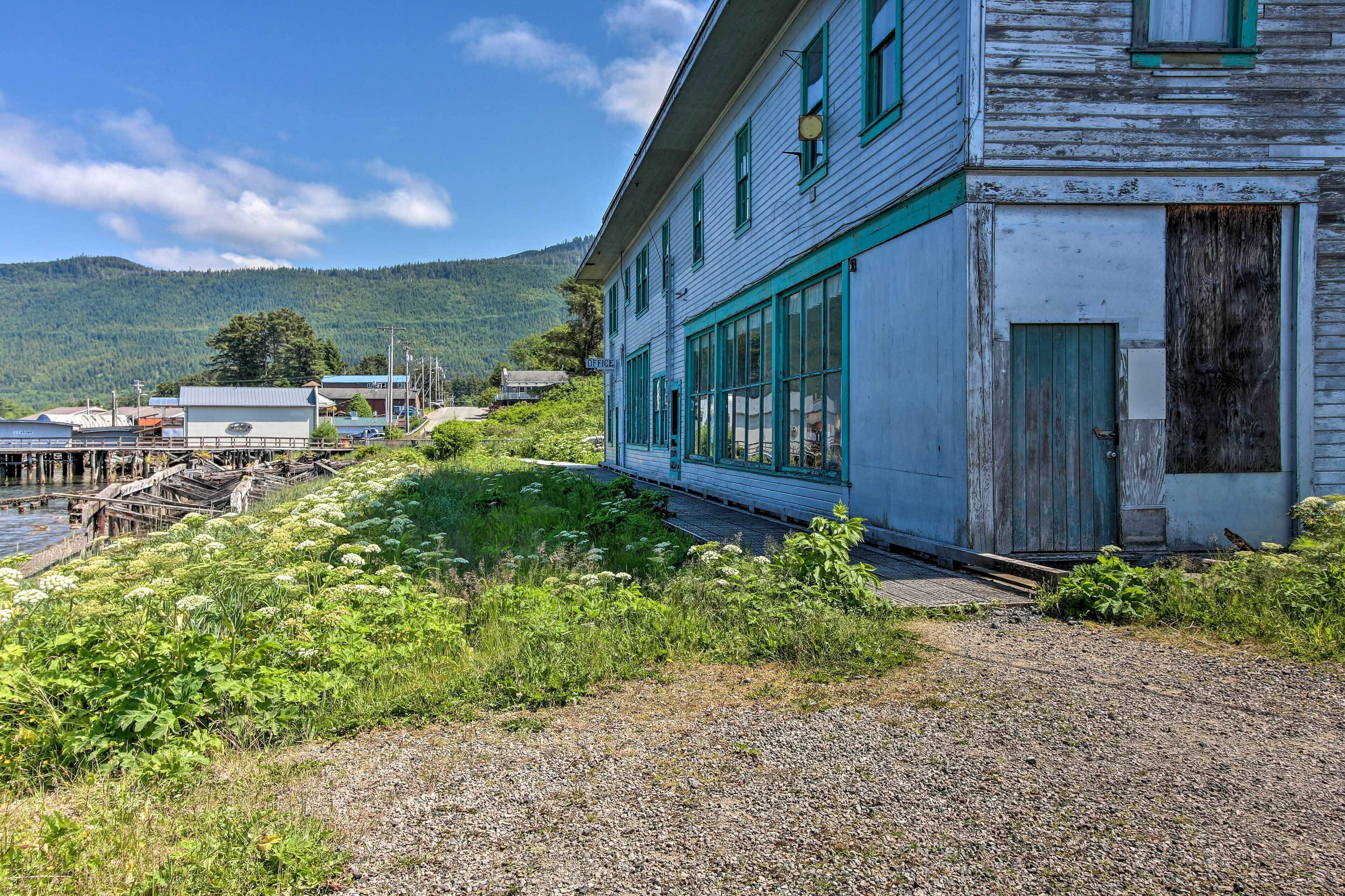 A rustic building with large windows and a wooden door, surrounded by greenery and a gravel path.