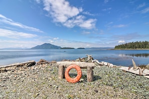 A rocky shore with driftwood and a tire, overlooking a calm lake with distant mountains and a forested island. - Waterfront Apt w/ Mtn View, Walk Into Town! (Craig)
