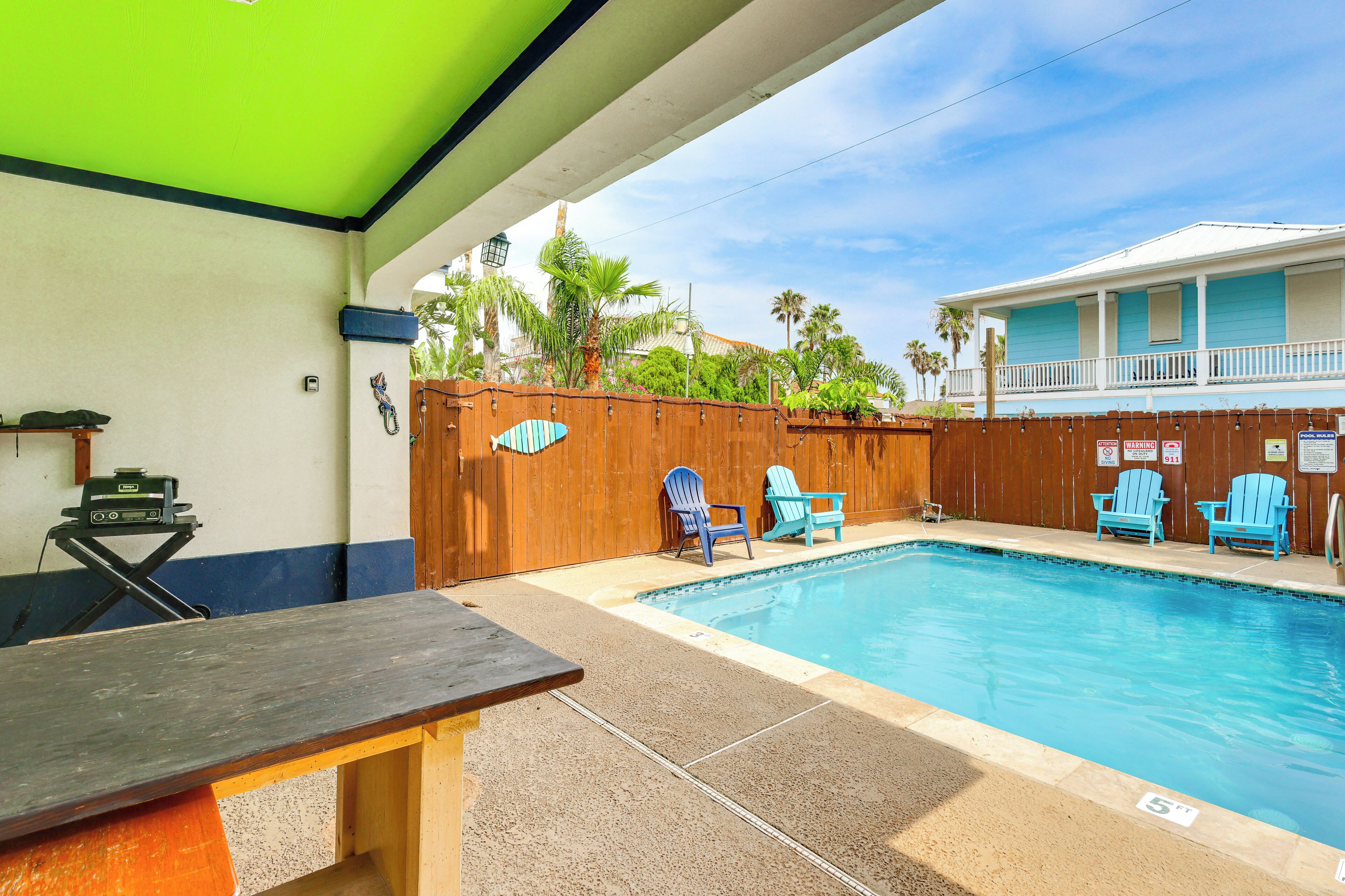 A swimming pool with blue chairs, a wooden fence, and a building with a balcony in the background.
