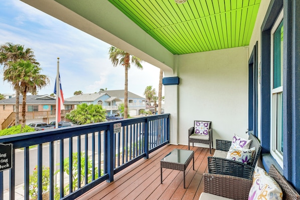 A balcony with a green ceiling, wicker furniture, and a view of palm trees and buildings.