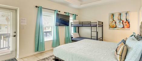 Une chambre avec un lit superposé, un lit simple, un ventilateur de plafond, une fenêtre avec des rideaux et un tableau représentant des guitares.