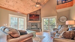 A spacious living room with a fireplace, leather sofas, and a large window. - Riverfront Tallulah Falls Cabin w/ Hot Tub! (Lakemont)