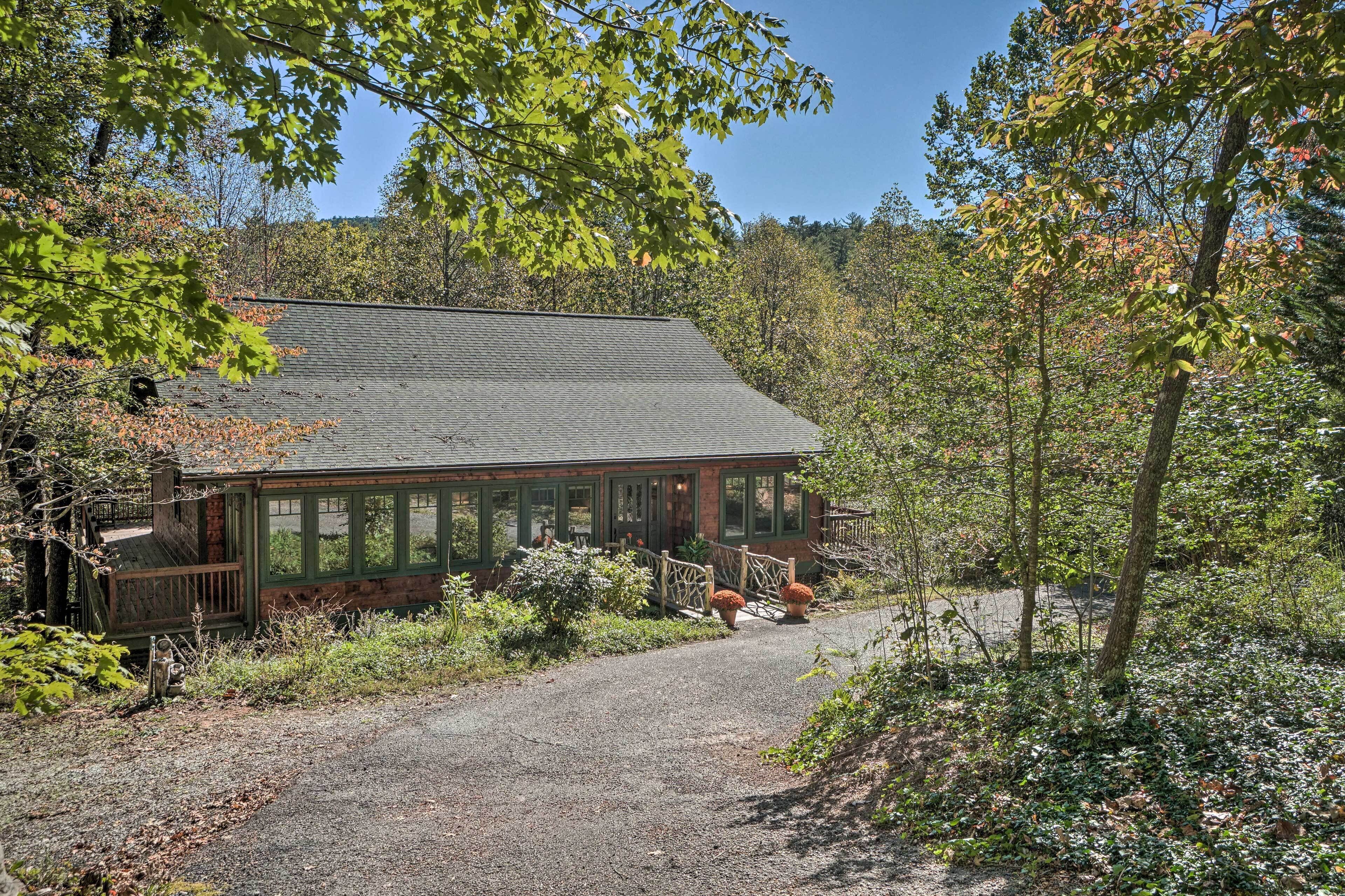 A rustic cabin with a gravel driveway, surrounded by dense forest and autumn foliage.
