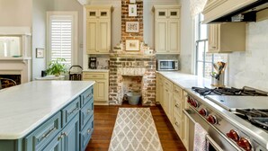 Una cocina con chimenea de ladrillo central, muebles de color claro, una isla grande y una estufa de acero inoxidable.