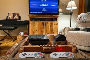 A living room with a television displaying a game menu, a wooden cabinet, a couch, and a tray with two game controllers. - Ellijay Cabin With Porch & Private Hot Tub! (Ellijay)