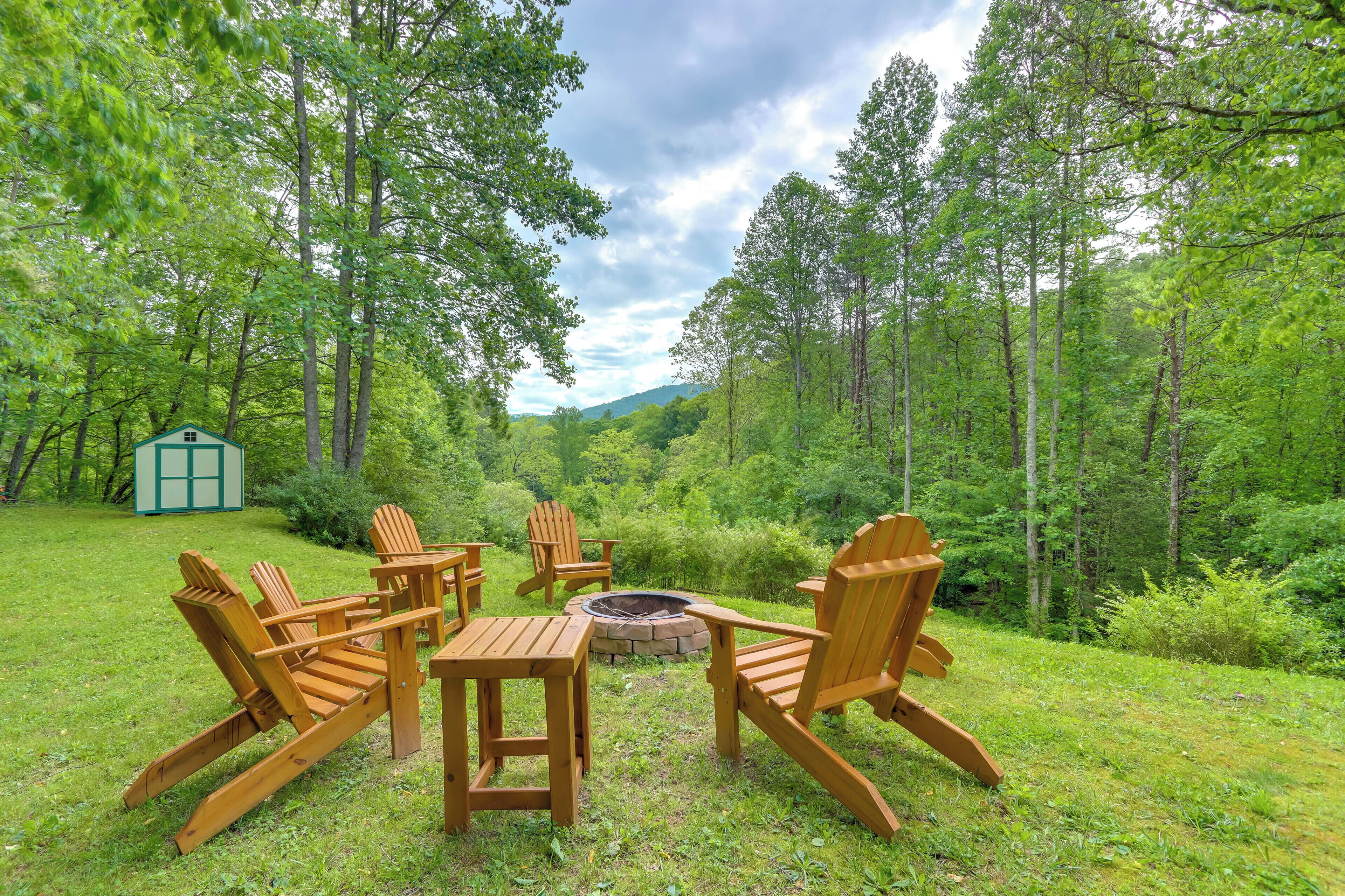 A wooden deck with Adirondack chairs and a fire pit surrounded by a lush forest.